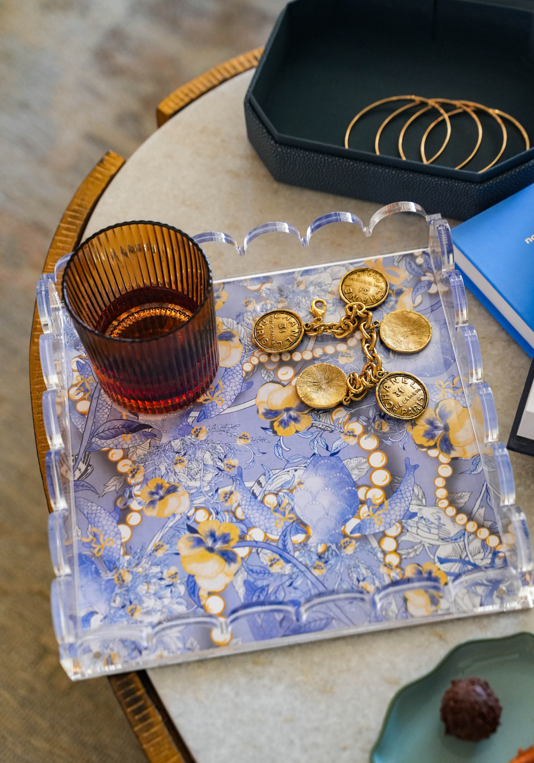 Clear acrylic tray with scalloped edges holding gold jewelry and amber glass on blue patterned surface.
