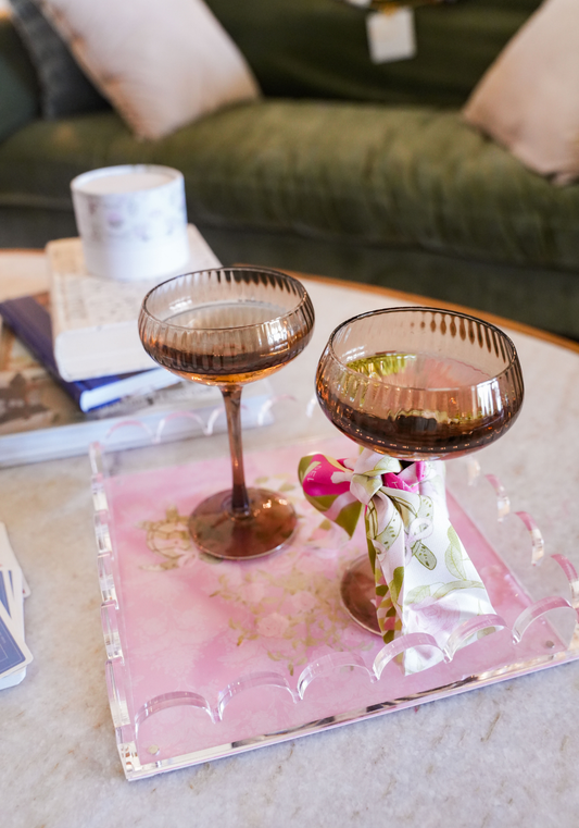 Pink acrylic tray with gold scalloped edges holding two amber coupe glasses on marble surface.