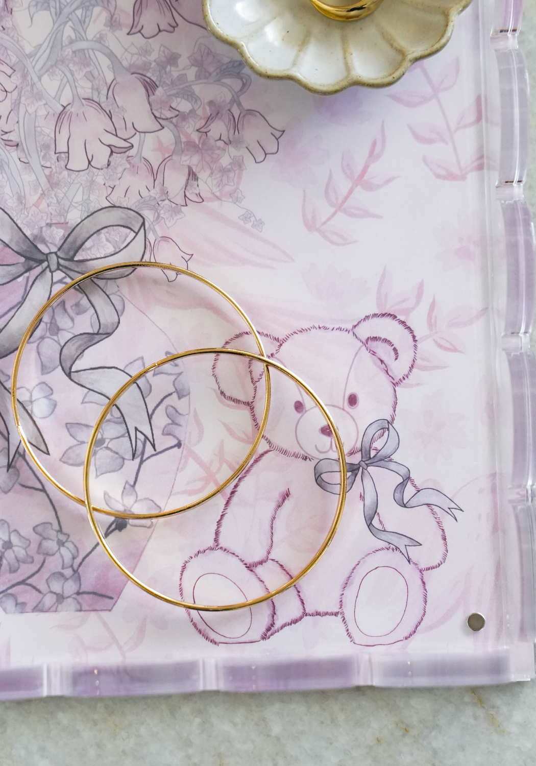 Clear acrylic tray with scalloped edges featuring a pink floral and teddy bear design and gold hoop earrings placed on top.