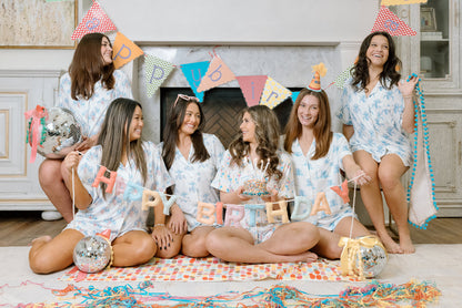 Six women wearing colorful birthday pajama shorts sets with party hats and birthday decorations indoors.