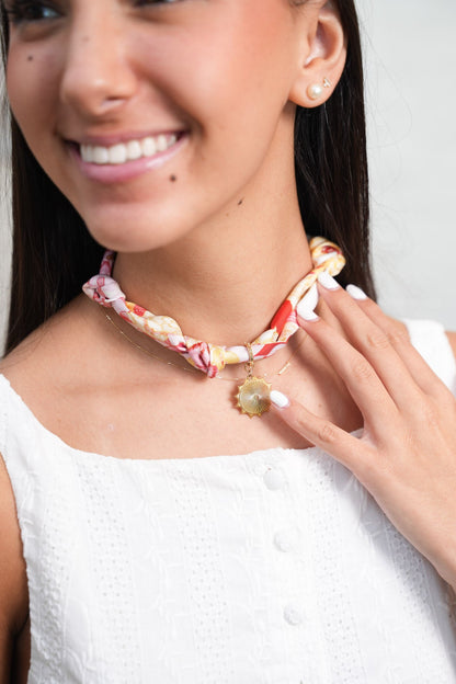 Light pink patterned scarflette with gold charm worn around the neck on a smiling woman in a white textured top.