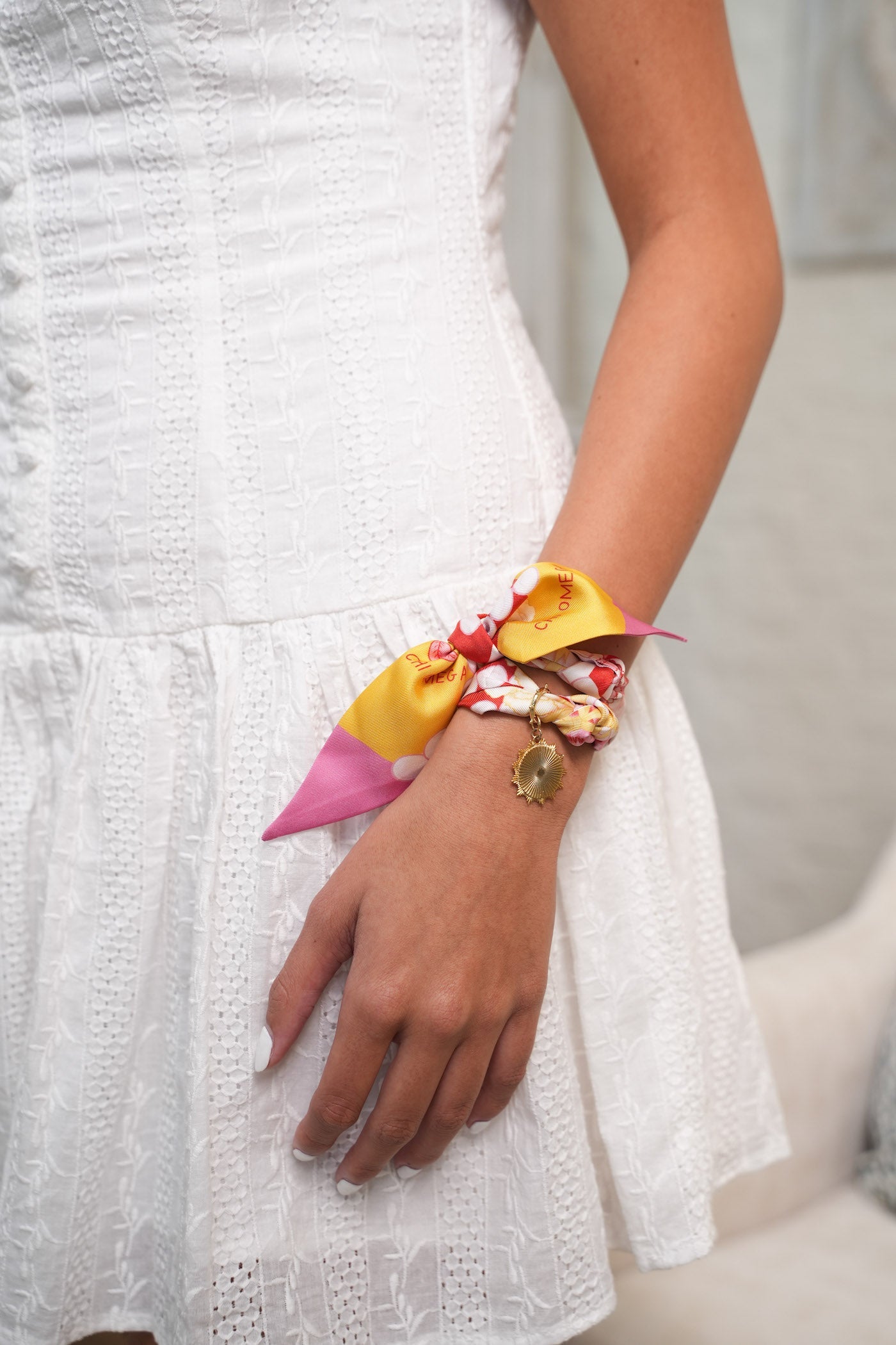 Silk Chi Omega scarflette in pink and gold tied around wrist with charm bracelet on woman in white dress.