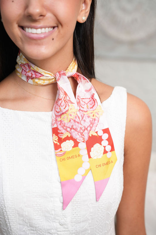 Pink, gold, and white Chi Omega scarflette tied around neck with floral and emblem patterns.